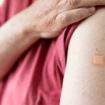 A man holds up his tshirt to reveal his upper arm with a band aid, indicating he has just received a vaccination