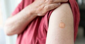 A man holds up his tshirt to reveal his upper arm with a band aid, indicating he has just received a vaccination