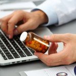 Male GP holds small jar of pills and types on laptop computer keyboard.